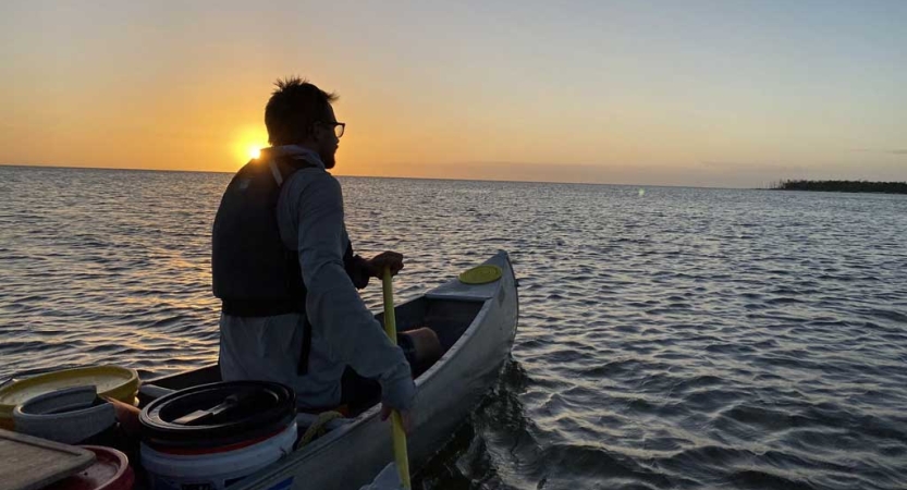 A person sits at the front of a canoe and paddles. The sun is behind them and appears to be setting. 